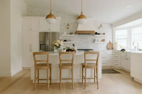 A bright, airy modern farmhouse kitchen with white subway tile, gold pendant lights, and a large island with three wooden stools.